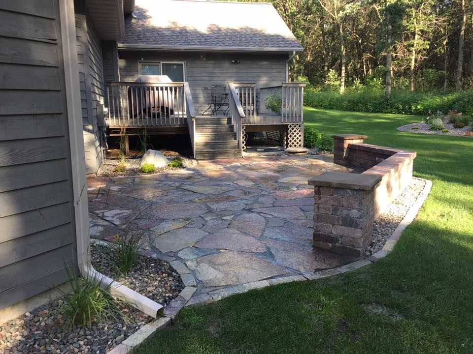 Stone patio and raised deck beside a house, with a low brick wall and grassy yard.