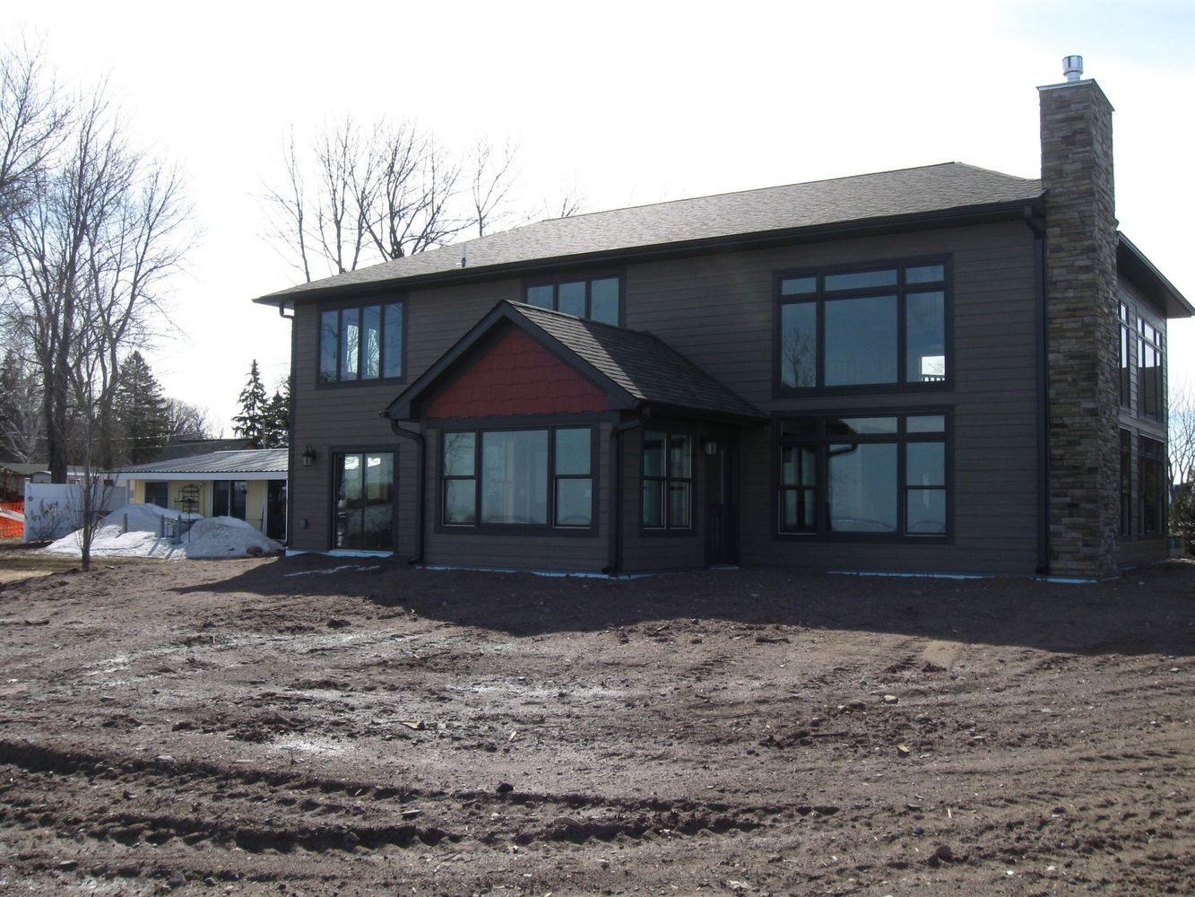 Two-story house with dark trim and stone chimney, set on a dirt lot under a clear sky.