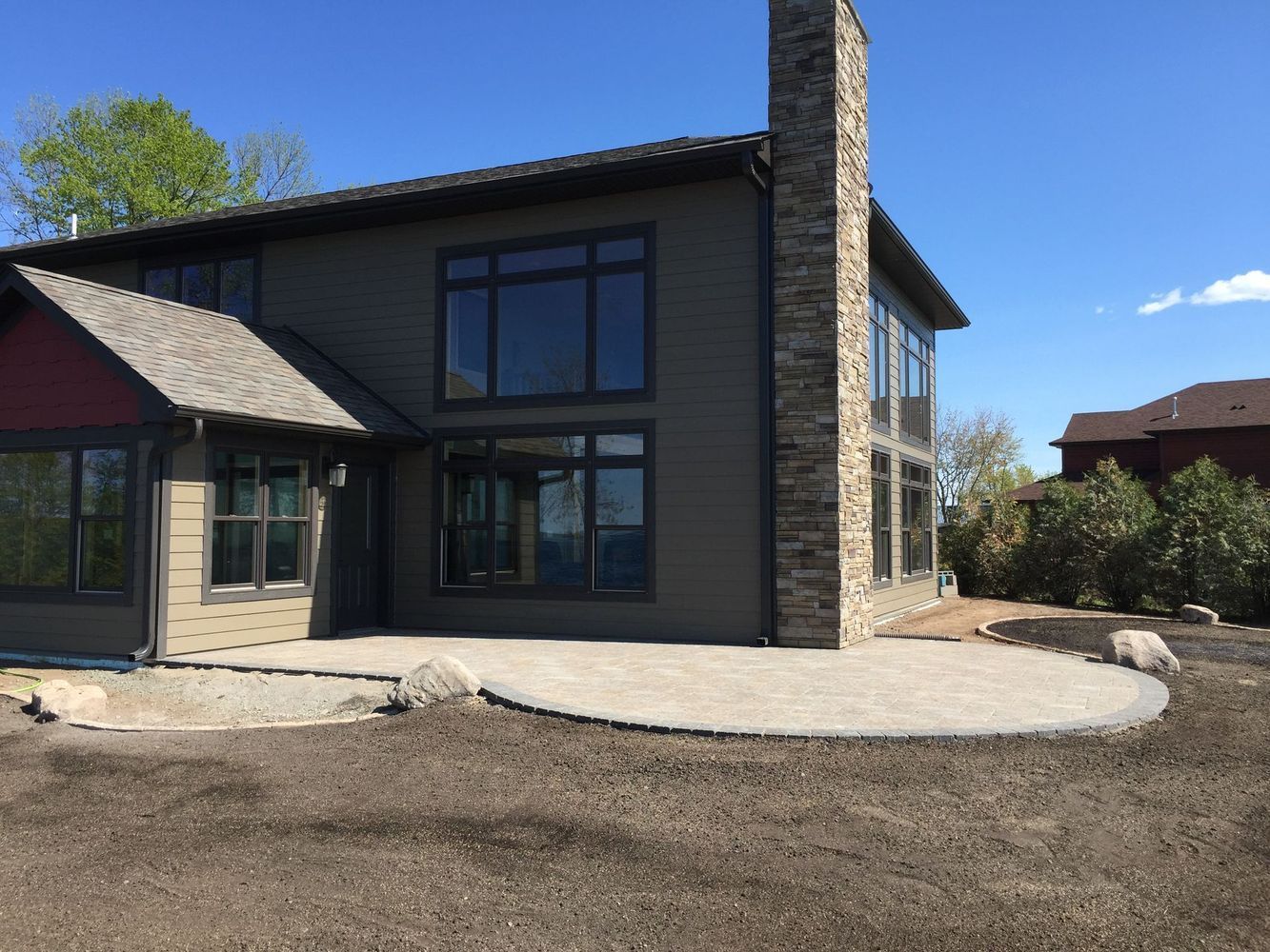 Modern two-story house with stone chimney, large windows, and a gravel driveway under a clear blue sky