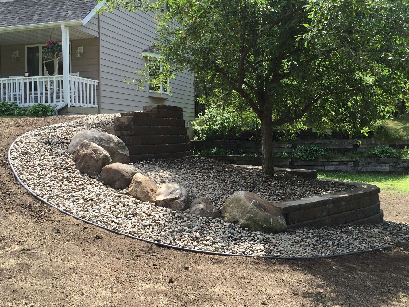 Curved rock-lined garden bed beside a house porch with a tree and stacked stone steps
