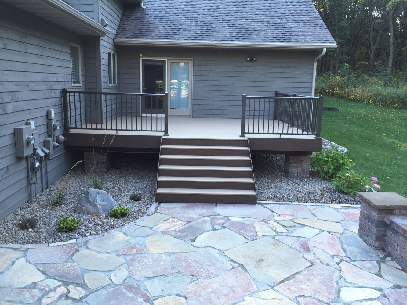 Stone patio with central stairs leading to a raised wooden deck and sliding glass door at the back of a house