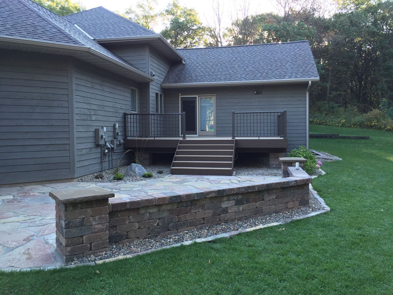 Backyard deck with steps, gray siding, stone retaining wall, and green lawn at dusk