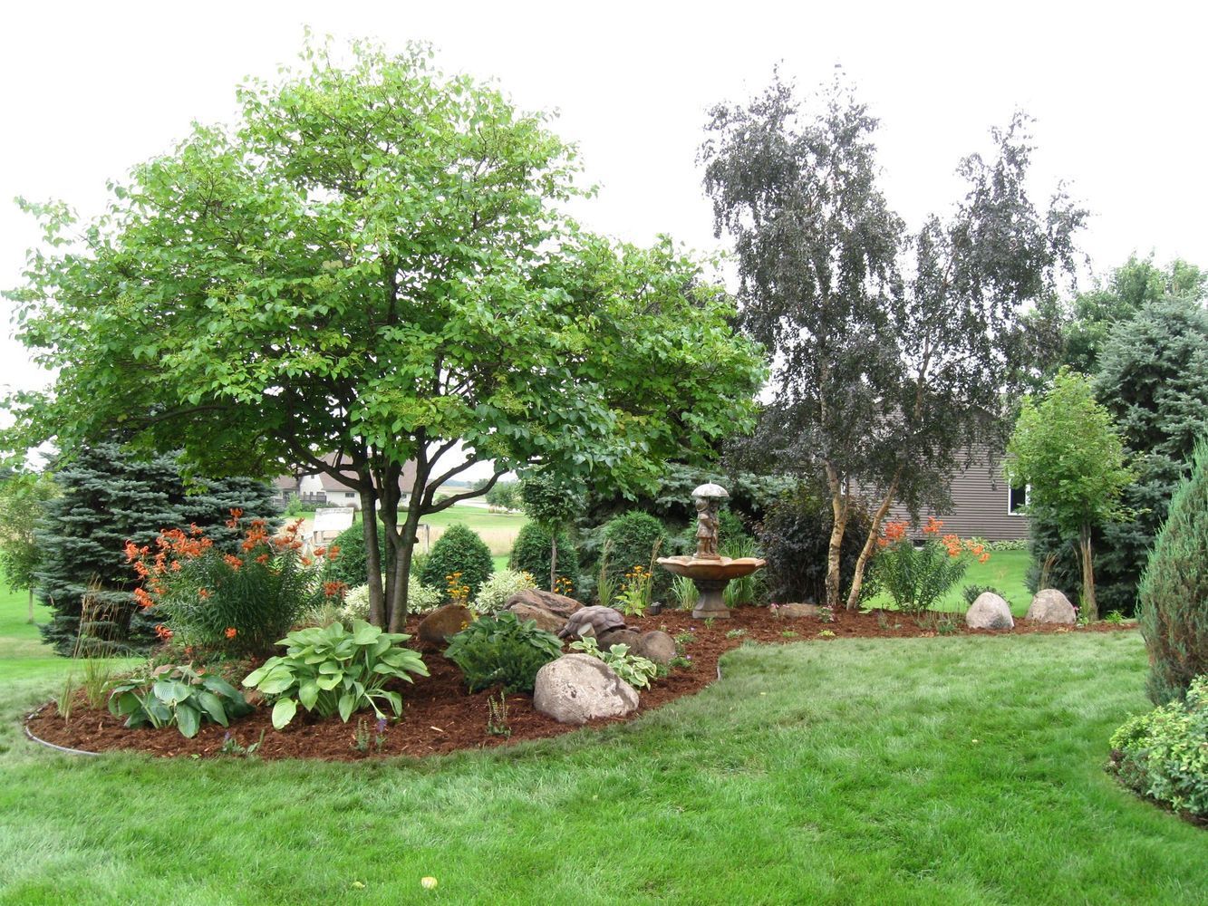 Lush backyard garden with leafy trees, shrubs, flowers, and a grassy lawn.