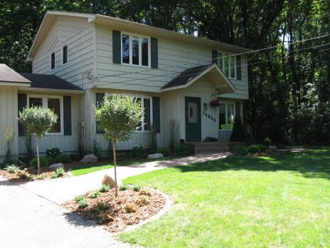 Two-story beige house with a front porch, driveway, and green lawn in a wooded neighborhood