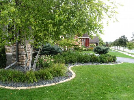 Landscaped yard with green lawn, trees, shrubs, and a stone-edged garden bed beside a sidewalk