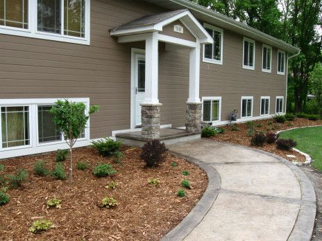Apartment entrance with white columns, curved sidewalk, and landscaped mulch beds under tan siding