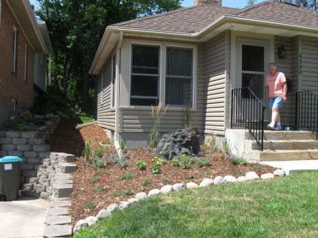 Woman standing on front steps of a beige house with a landscaped yard and stone border