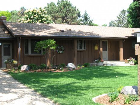 Single-story brown house with a green lawn, shrubs, and a driveway under a sunny sky