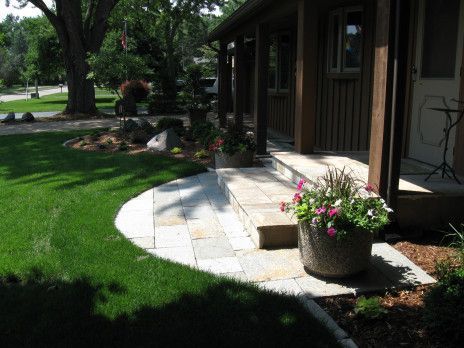 Curved stone patio beside a house, with a potted flower arrangement and lush green lawn.
