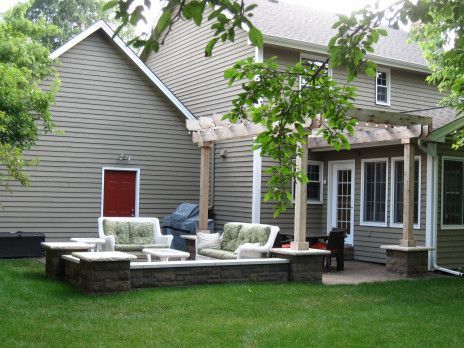 Backyard patio with white cushioned chairs and stone seating beside a gray house and green lawn