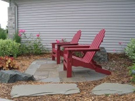 Two red Adirondack chairs on a stone patio beside a gray house wall and garden bed.