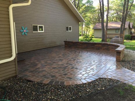 Wet brick patio beside a house, with a low retaining wall and trees in the background.