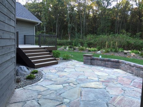 Stone patio with steps beside a house, bordered by a low wall and trees in the background
