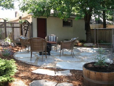 Patio with wicker chairs and table under a tree beside a small shed and garden beds