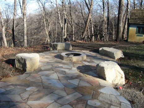 Stone patio with a circular fire pit and large boulders in a wooded backyard