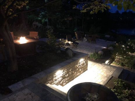 Dim garden patio at night with a lit fire pit, stone wall, and outdoor seating.