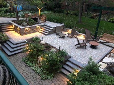 Tiered backyard patio with stone steps, built-in lighting, seating, and garden beds at dusk