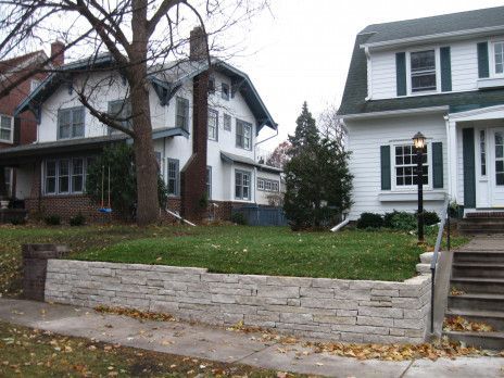 Two white houses with porches and a stone retaining wall along a tree-lined street in autumn