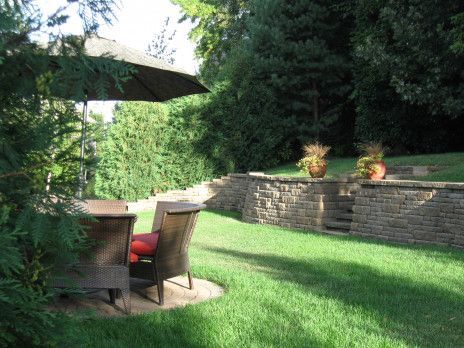 Patio with table and chairs under umbrella beside a stone retaining wall and lush green lawn.