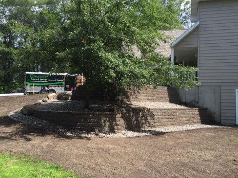 Large tree in a yard beside a gray house, with a paved path and a truck in the background.