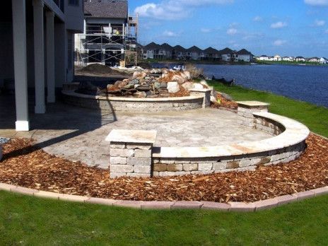 Curved stone patio beside a waterfront lawn, with houses and blue water in the background