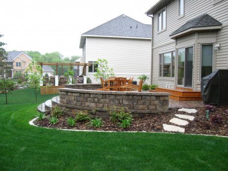 Backyard patio with stone retaining wall, wooden deck, and white houses in the background.