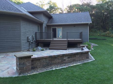 Backyard deck and patio beside a gray house, with stone retaining wall and green lawn