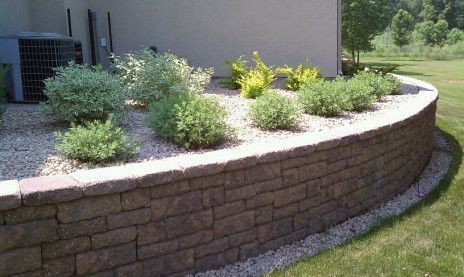 Curved stone retaining wall with landscaped shrubs beside a house and lawn