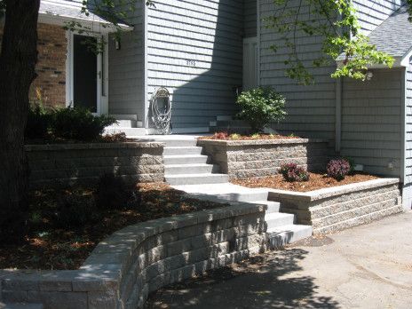 Front steps and stone retaining walls leading to a gray house with a driveway and shrubs