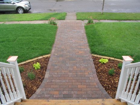 Brick walkway leading from a porch to the street, bordered by green lawn and small garden beds