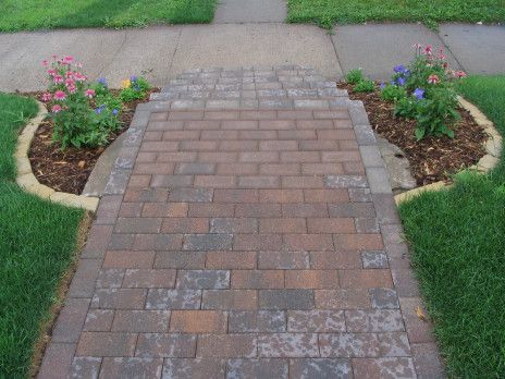 Brick walkway leading to a sidewalk, bordered by mulch beds with flowers and green lawn