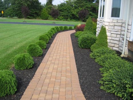 Brick walkway beside a stone house, bordered by trimmed green shrubs and a lawn