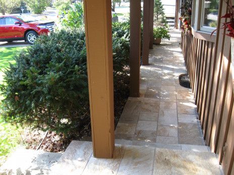 Covered outdoor walkway with wooden posts, stone path, shrubs, and a red car in the background