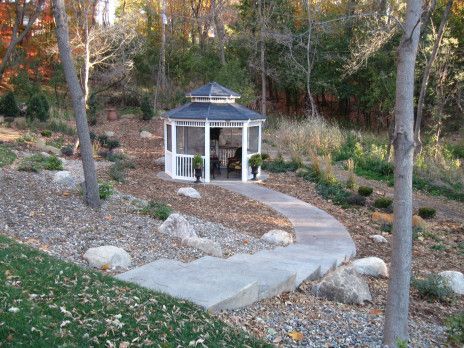 Small white gazebo beside a curved concrete path in a wooded garden with rocks and fall foliage