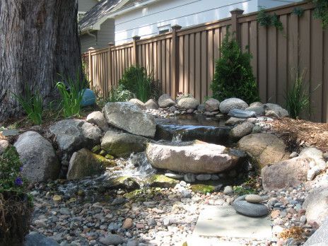 Small backyard rock waterfall with flowing water, pebbles, and a wooden fence behind it