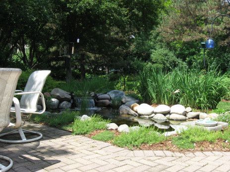 Backyard pond with rocks and greenery beside patio chairs and brick pavers