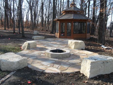 Stone fire pit with benches in a wooded park beside a small wooden pavilion.