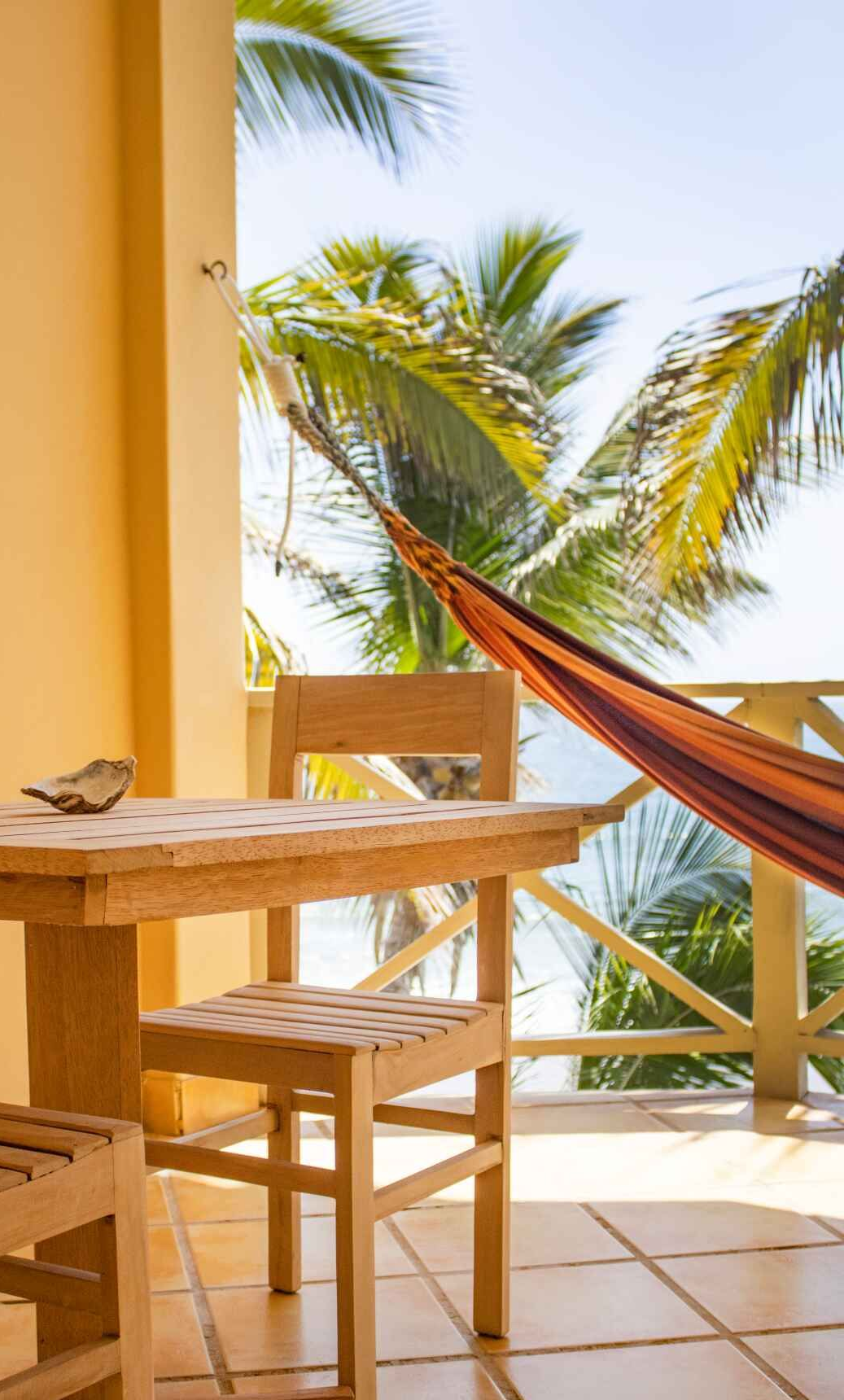 A table and chairs on a balcony with a hammock overlooking the ocean.