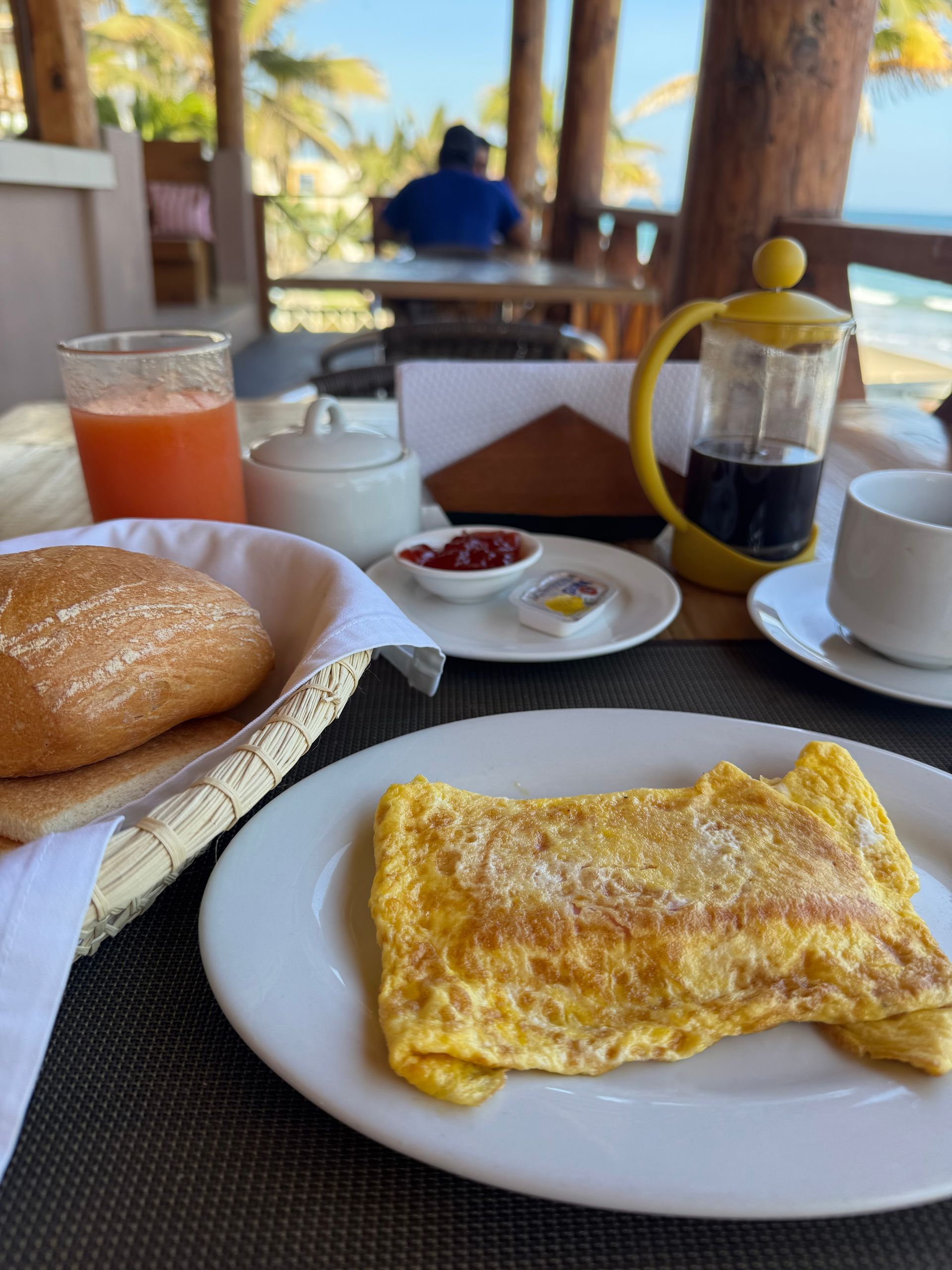 Breakfast at a beachside restaurant: omelet, bread, juice, coffee, person in background.