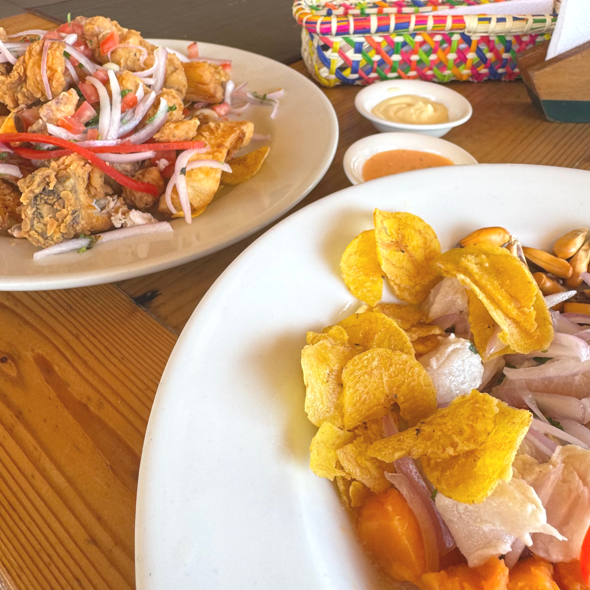 Two plates of food on a wooden table: fried fish with onions and a ceviche dish with plantain chips.