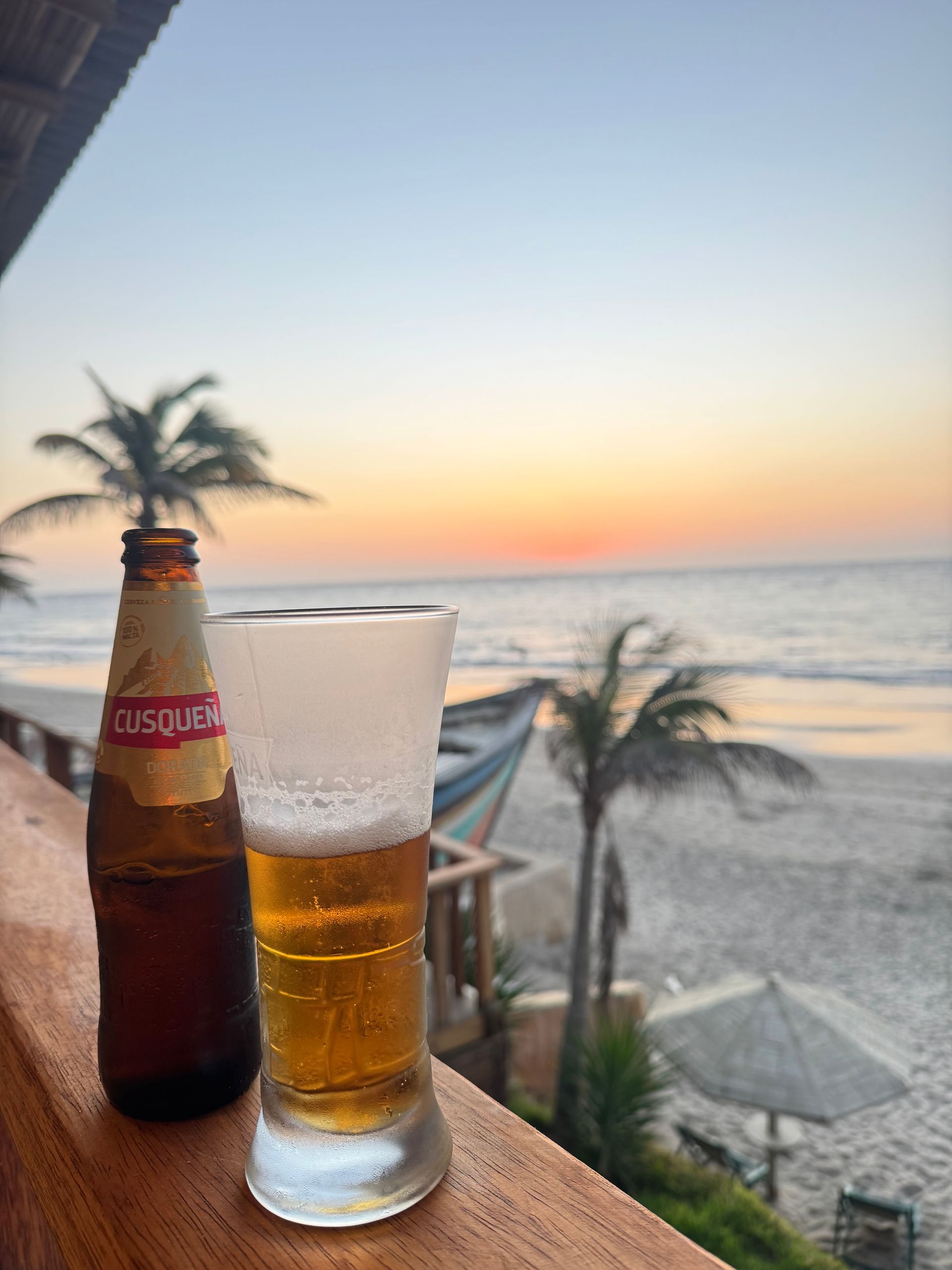 Beer bottle and glass on a railing overlooking a beach at sunset.