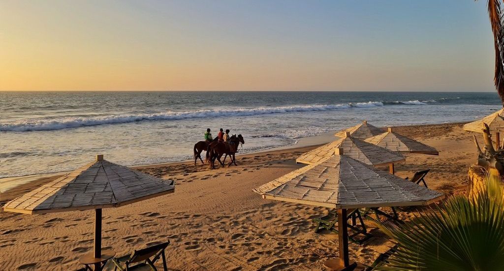 Beach scene with horses being ridden near the water; umbrellas on sand. Sunset colors.