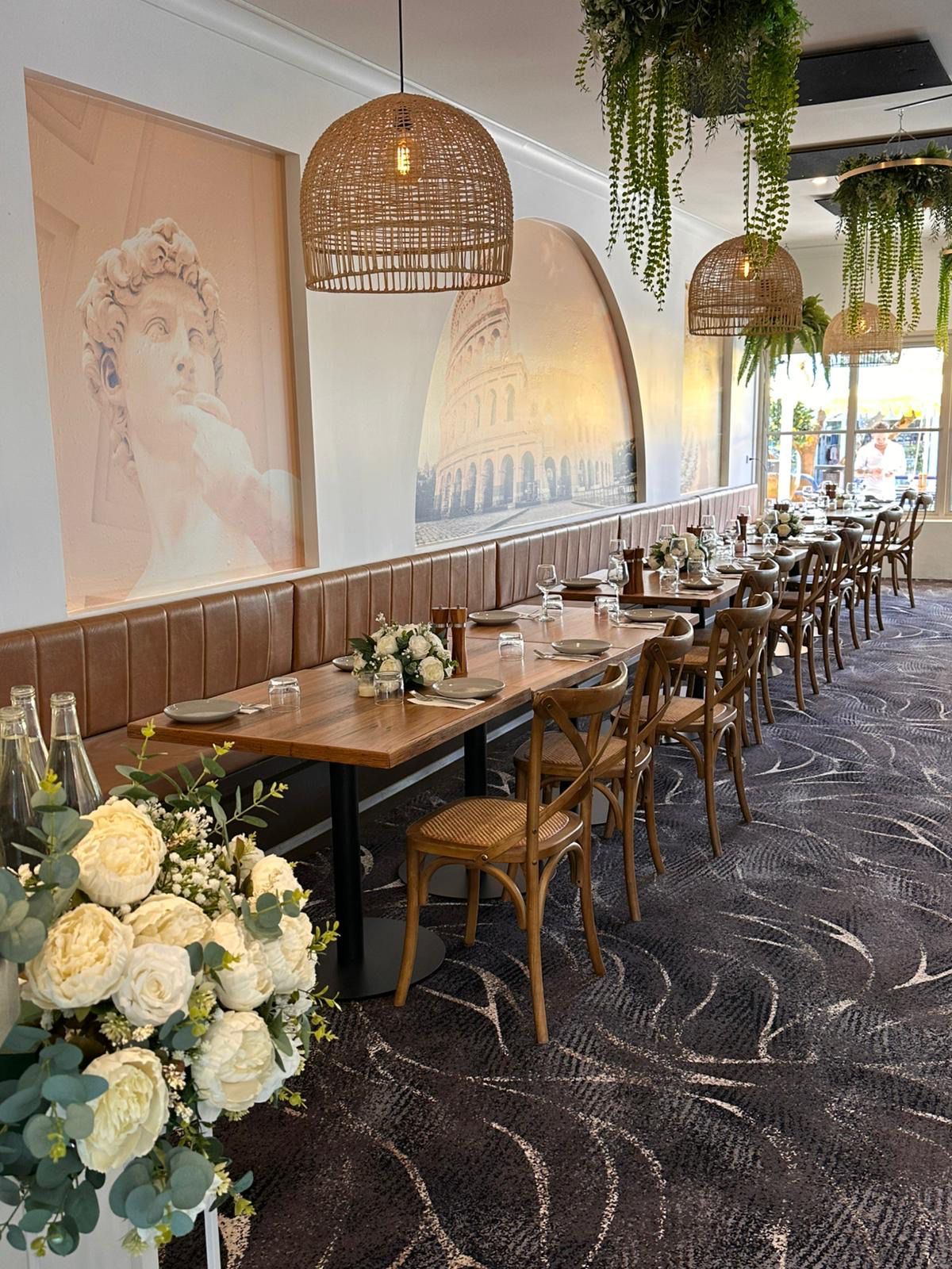 Restaurant interior with long wooden table, leather bench, woven light fixtures, and floral arrangement — Pizza in Huskisson, NSW