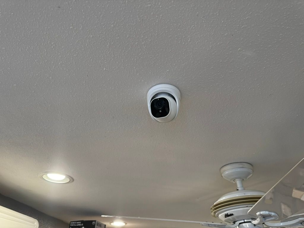 A security camera is mounted on the ceiling of a room next to a ceiling fan.