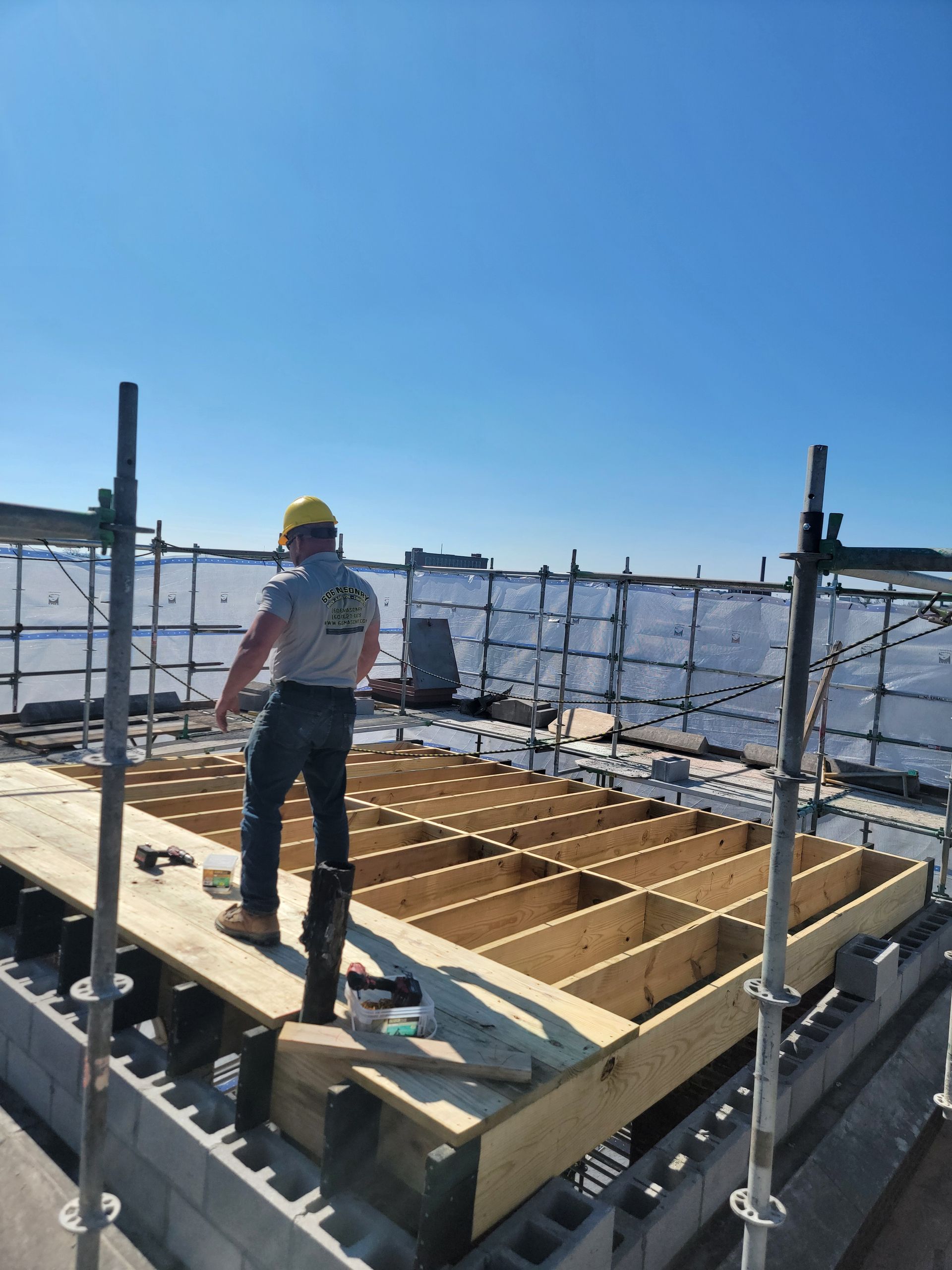 Worker on a rooftop wooden frame under construction, wearing a yellow hard hat against a clear blue sky.