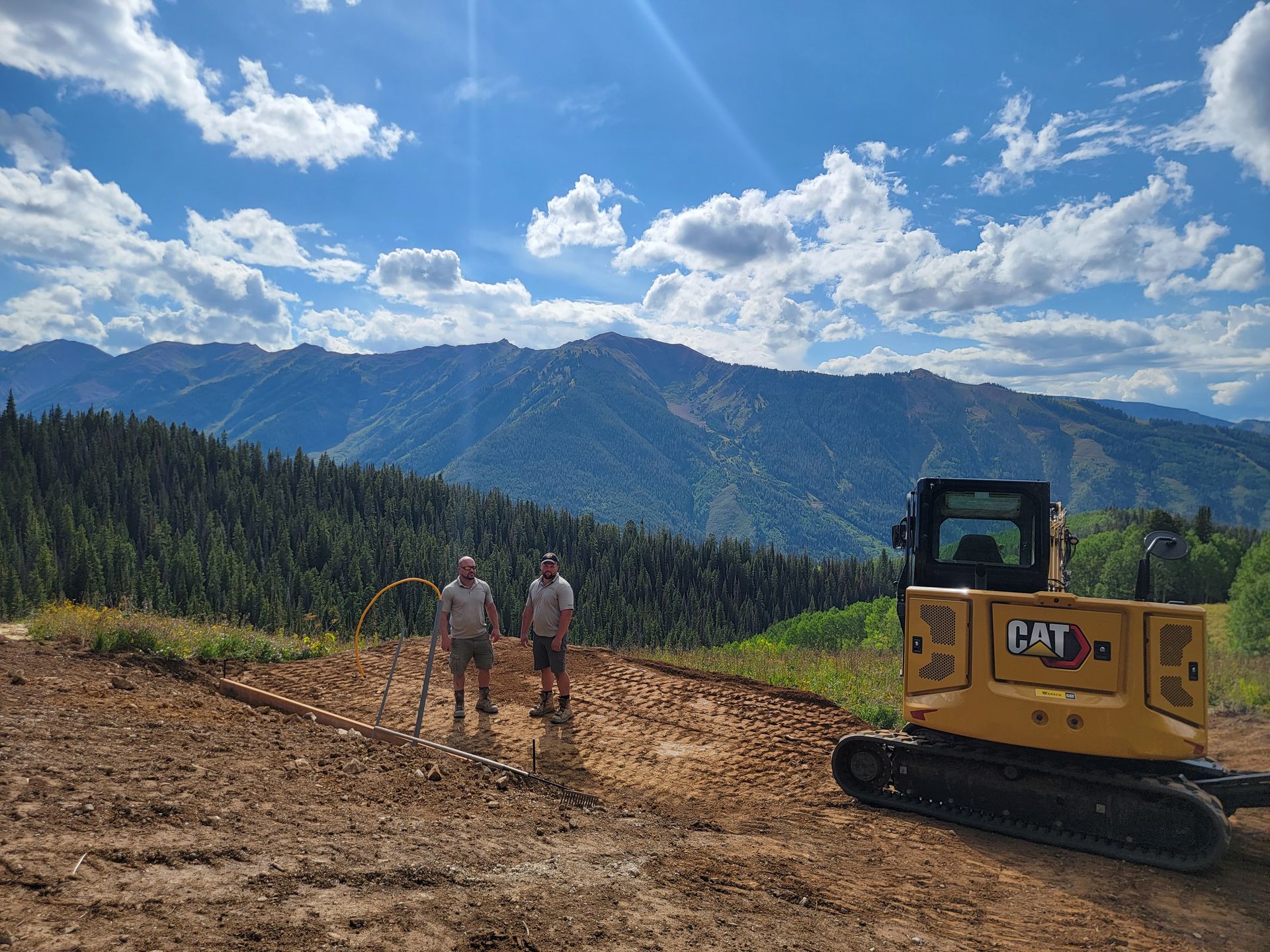 Two workers and a bulldozer on a dirt site with mountains and blue sky in the background