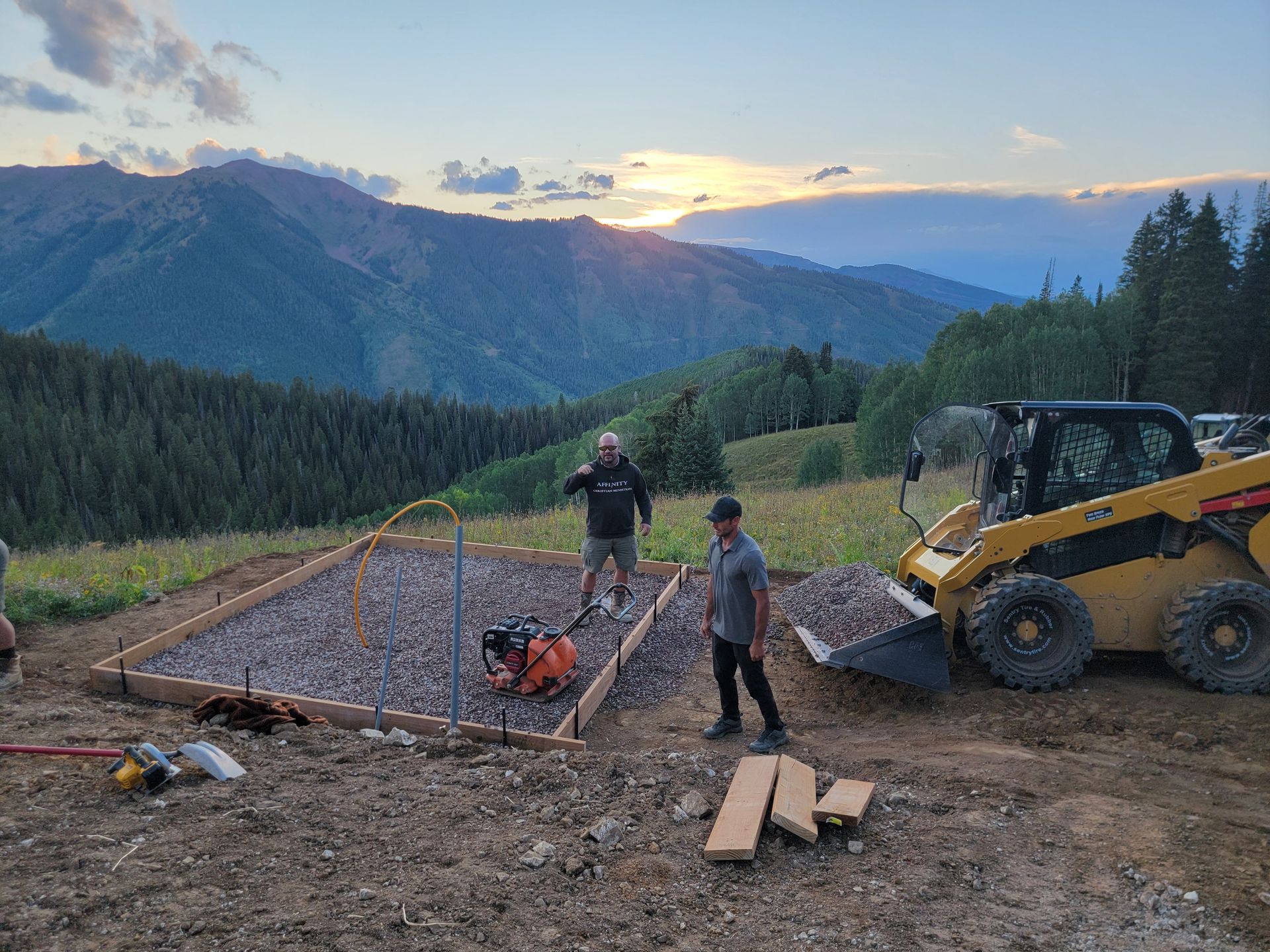 Workers building a wooden platform on a mountain ridge at sunset, with a skid-steer loader nearby.