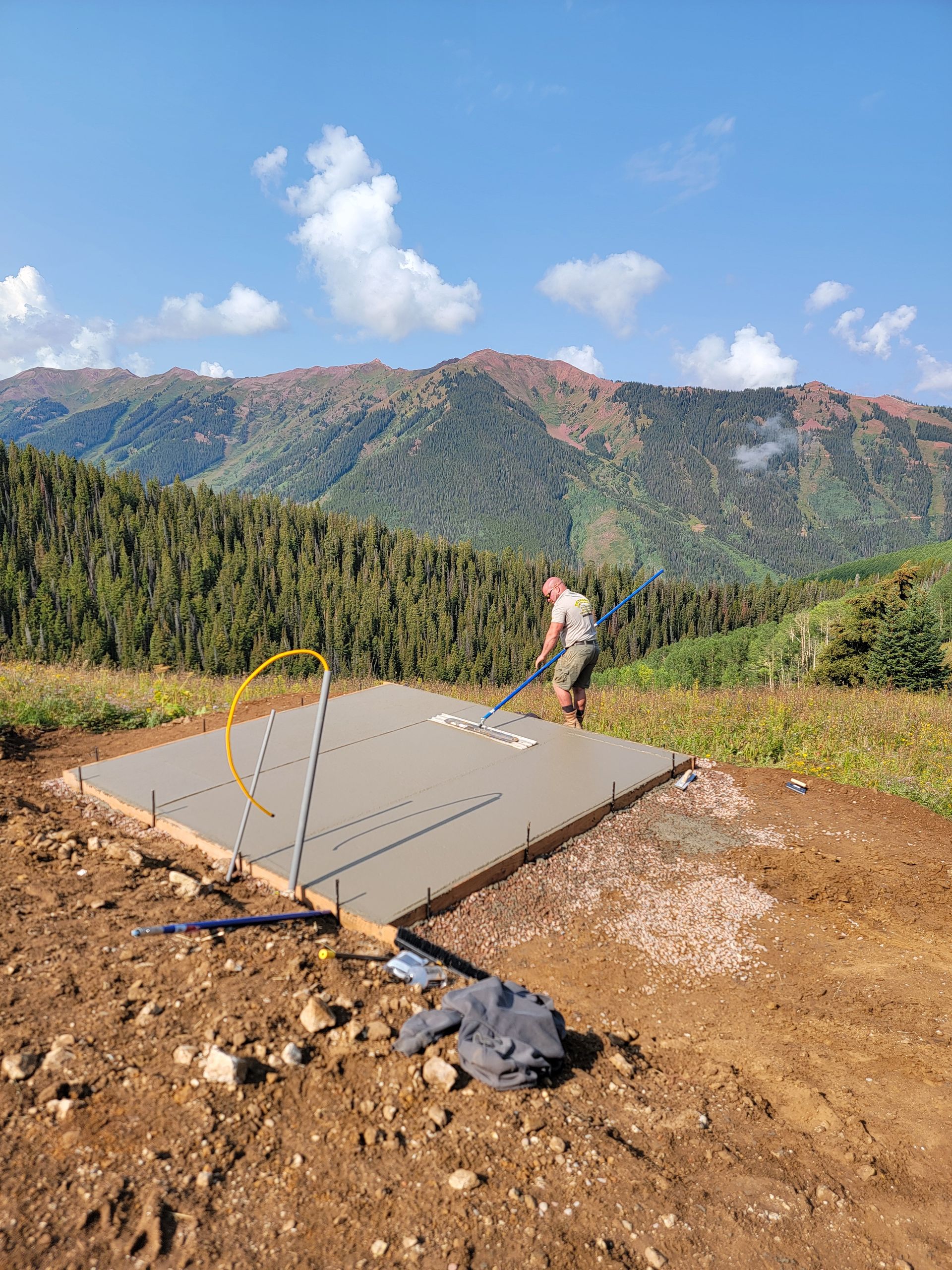 Person standing on a concrete pad in a dirt field with mountains and blue sky in the background