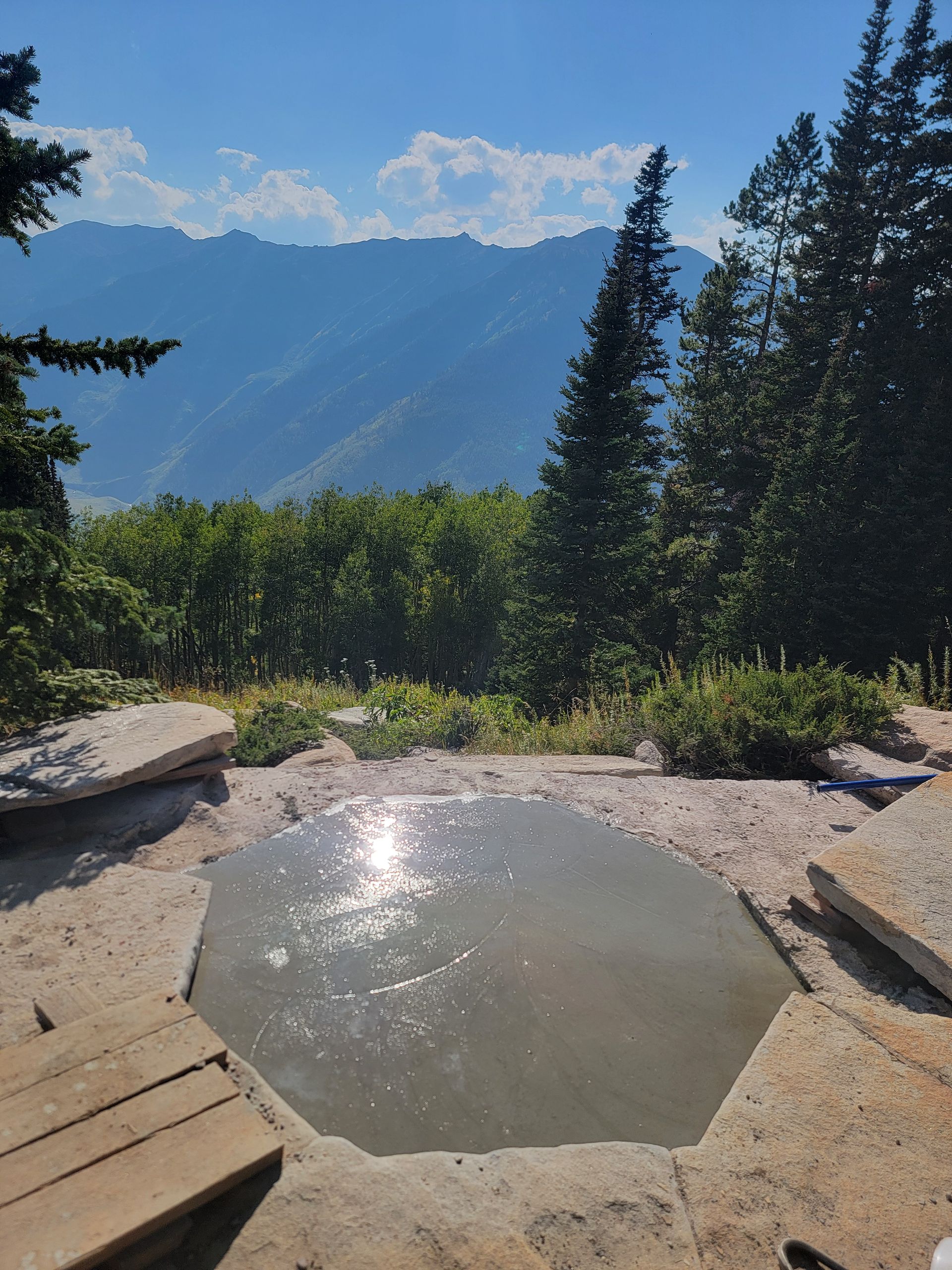 Small stone hot spring pool with mountains and evergreen trees in the background.