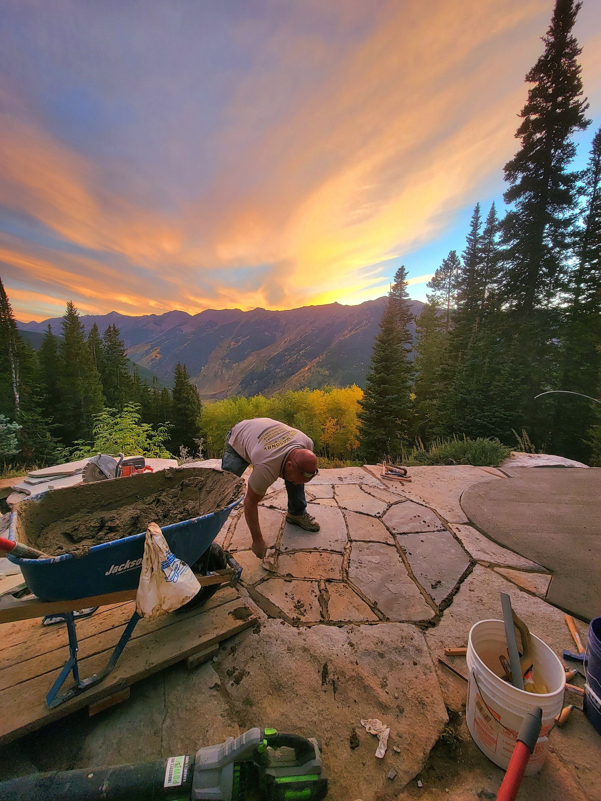 Person bending over patio stones beside a wheelbarrow at sunset, with mountains and pine trees in the background